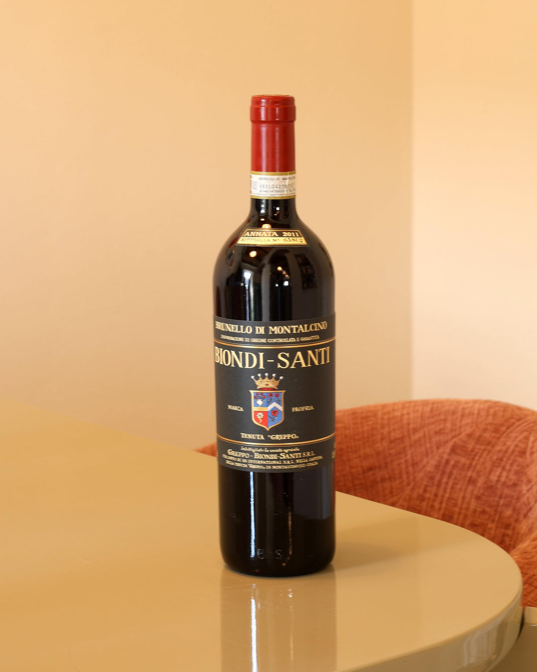 Bottiglia di Biondi Santi Brunello di Montalcino 2011 con etichetta nera classica e capsula rossa su tavolo beige.