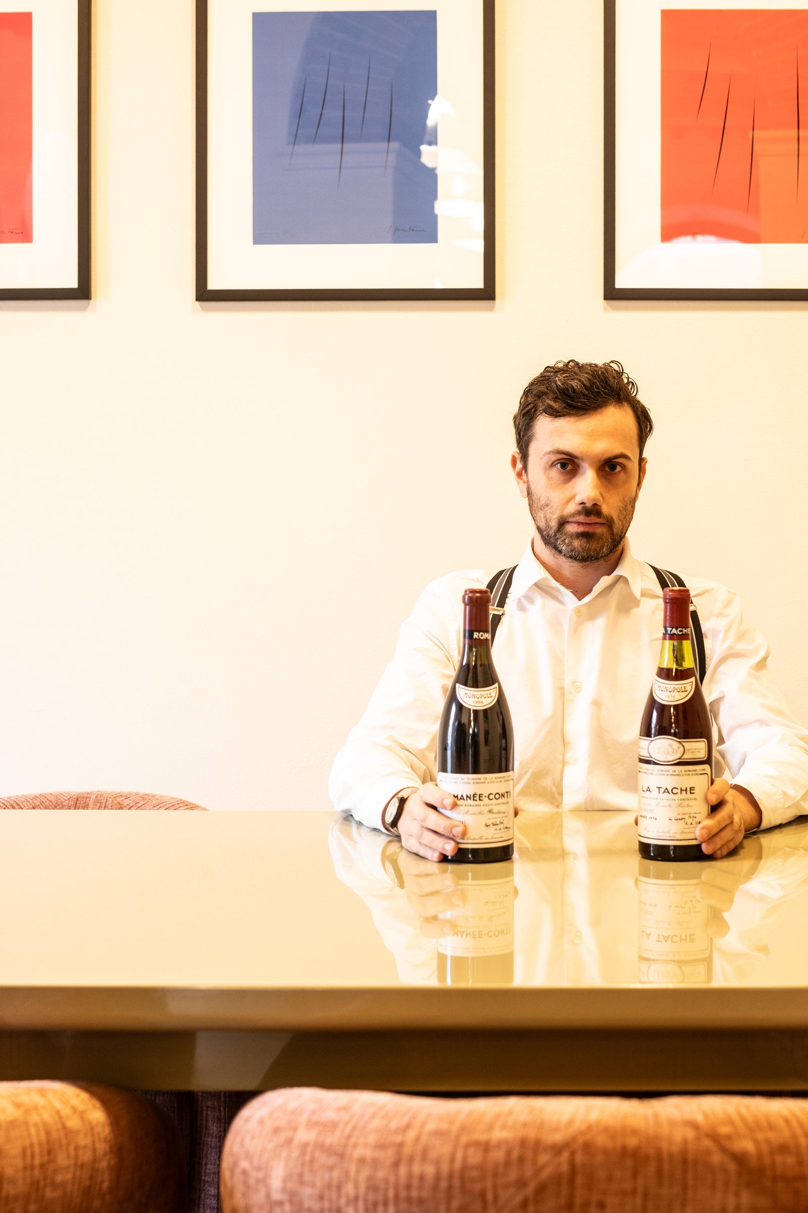 Man in a white shirt and suspenders sits at a table holding two vintage bottles of Romanée-Conti and La Tâche wine.