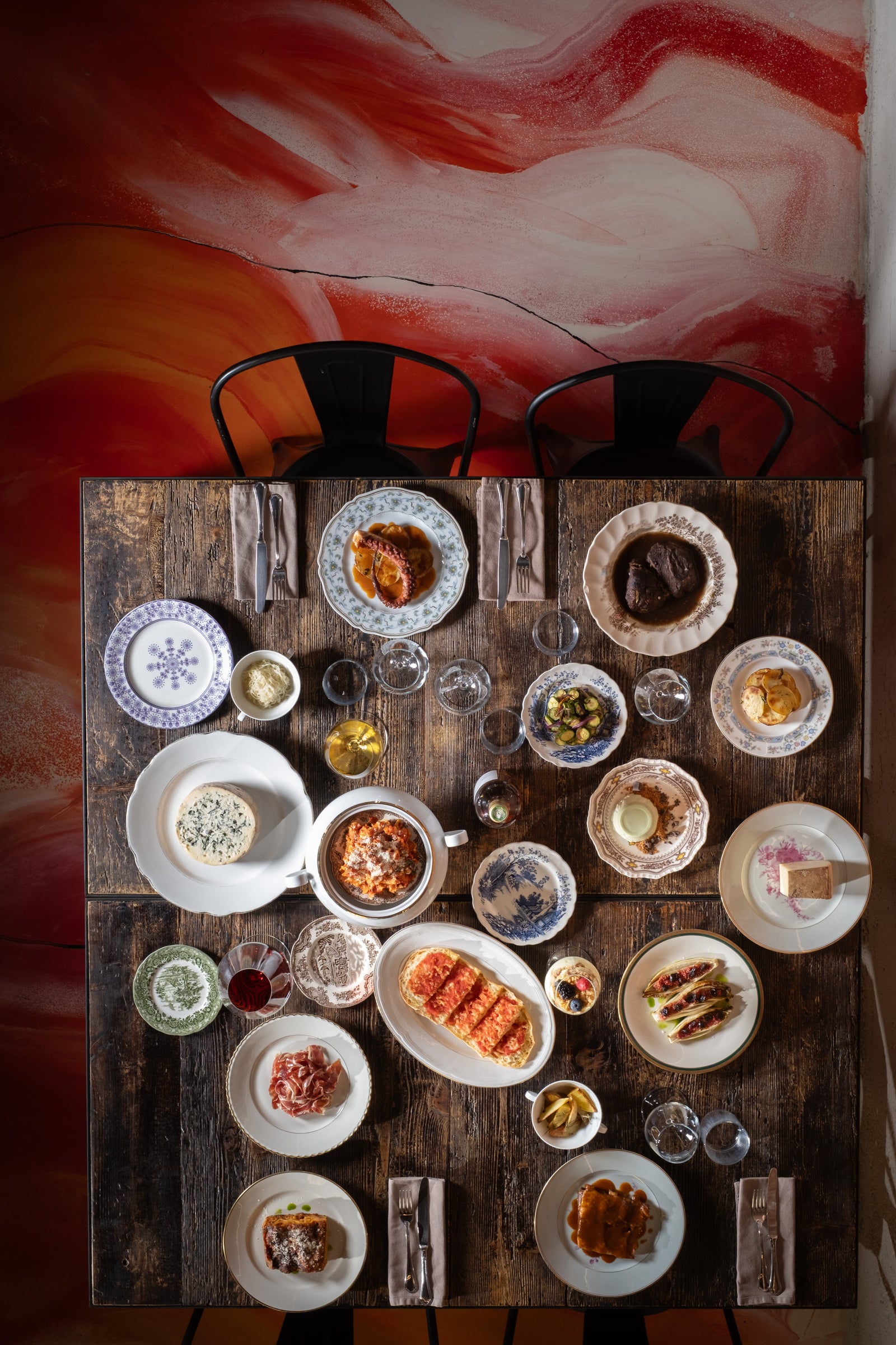 Overhead view of a rustic wooden table filled with various gourmet tapas, small plates, and wine glasses on a vibrant red wall background.