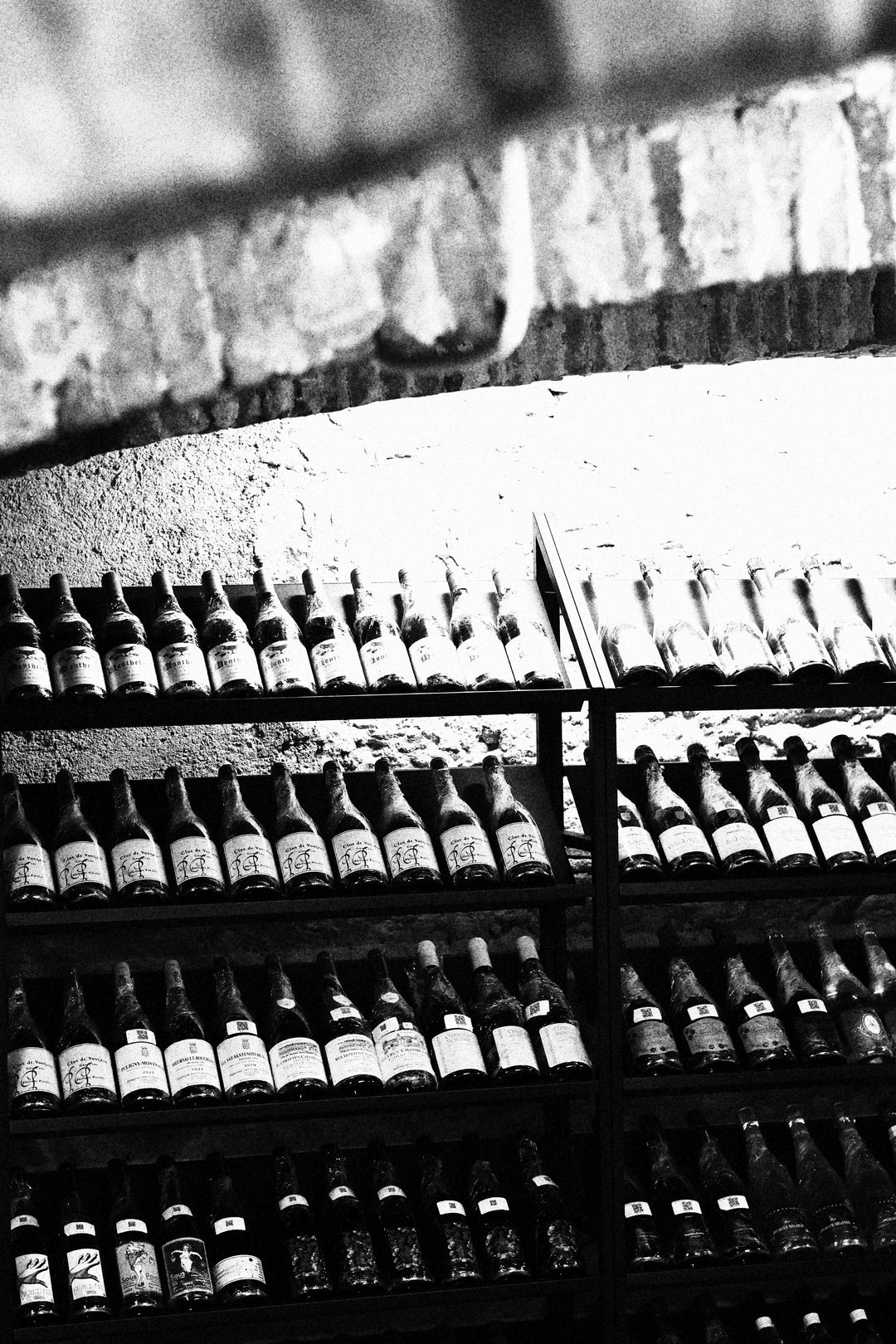 Black and white view of wine bottles neatly arranged on dark metal shelves in a stone cellar.