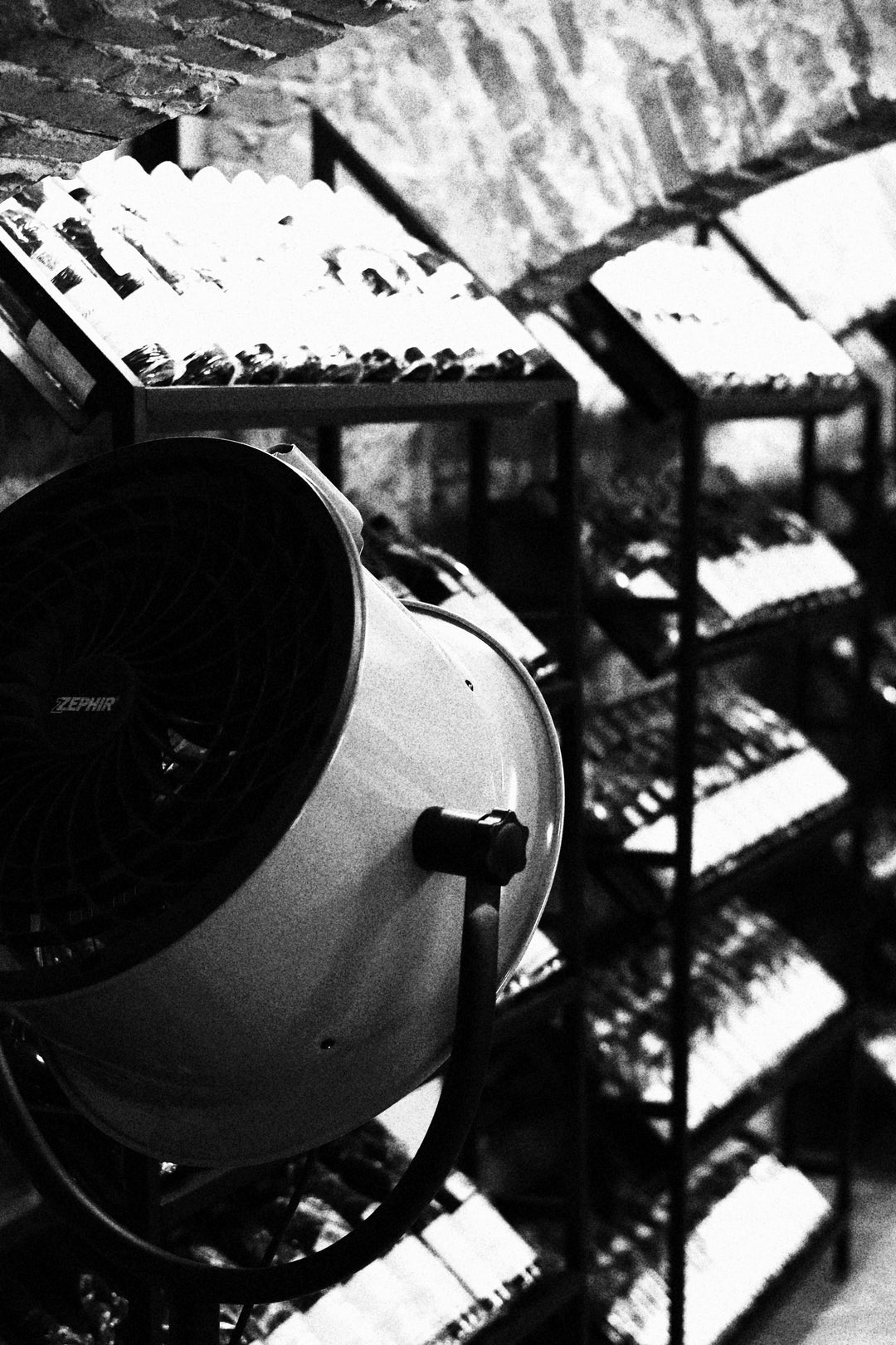 A Zephir fan positioned in front of metal wine racks inside a rustic, arched stone cellar in black and white.