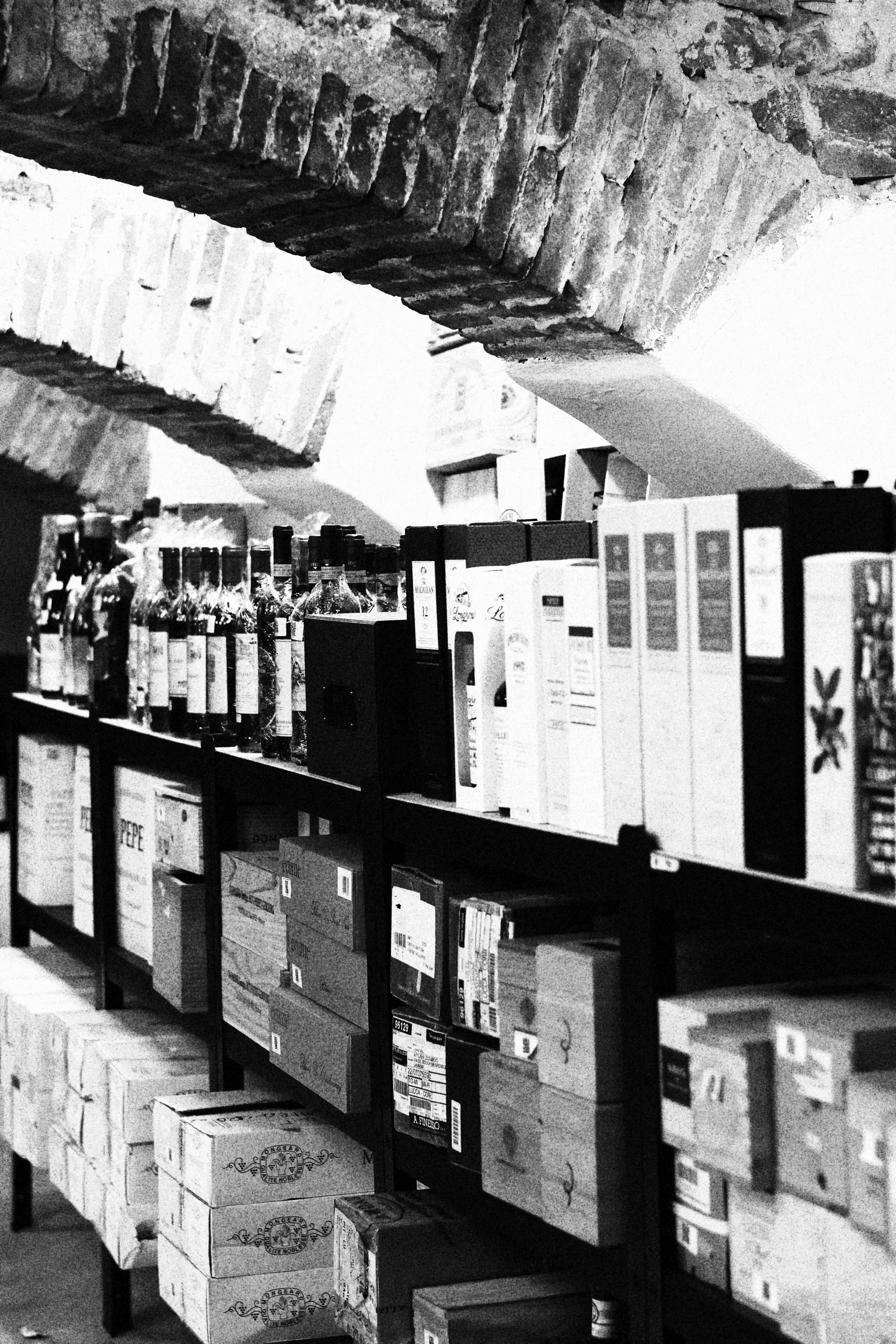 Black and white view of wine bottles and cardboard boxes arranged on dark metal shelves under a rustic brick archway.