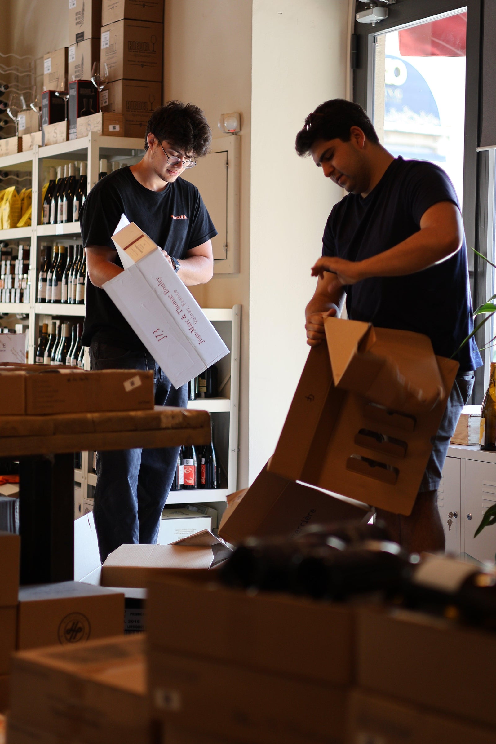 Two men in a wine shop unpacking cardboard boxes of wine bottles near shelves stocked with glass bottles.