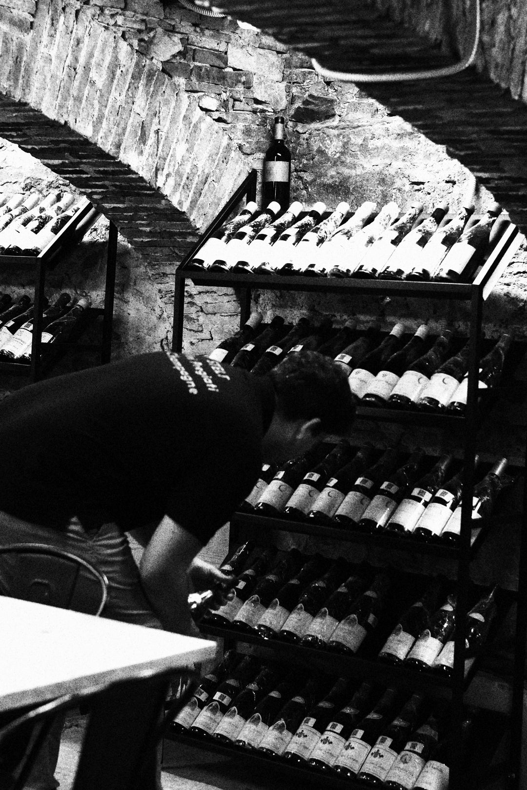 Person selecting a wine bottle from metal racks in a rustic stone cellar with arched brick ceilings.