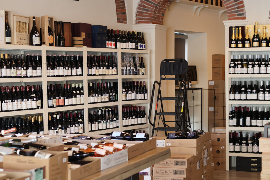 Shelves filled with various wine bottles in a shop, featuring a black folding ladder and cardboard shipping boxes.
