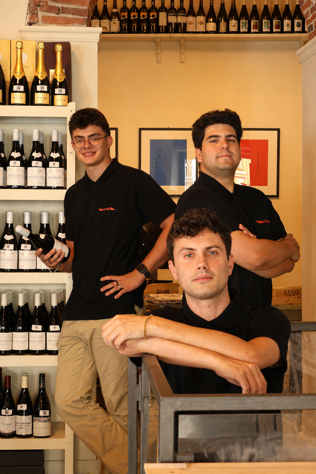 Three young men in black polo shirts posing in a wine shop with shelves of bottles in the background.