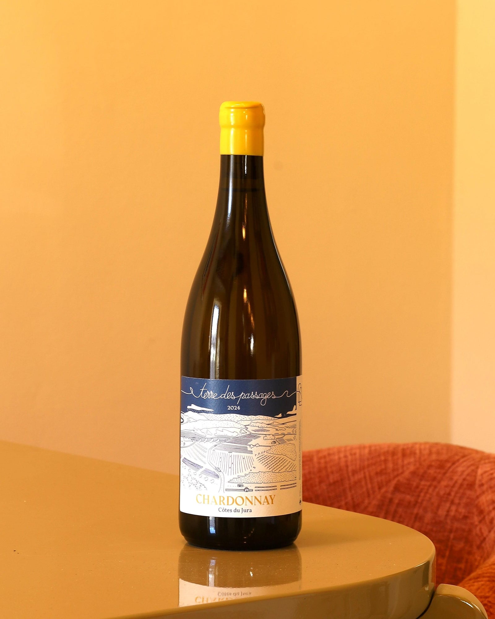 Bottiglia di vino bianco Terre des Passages Chardonnay 2024 con etichetta illustrata blu e capsula gialla su tavolo.