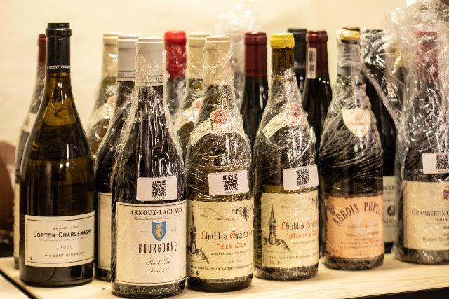 Assorted French wine bottles with labels and QR codes, many wrapped in protective plastic film on a wooden shelf.