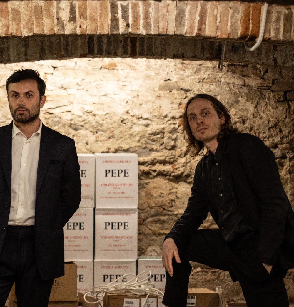 Two men in dark suits standing in a rustic stone wine cellar next to stacked boxes labeled Azienda Agricola Pepe.
