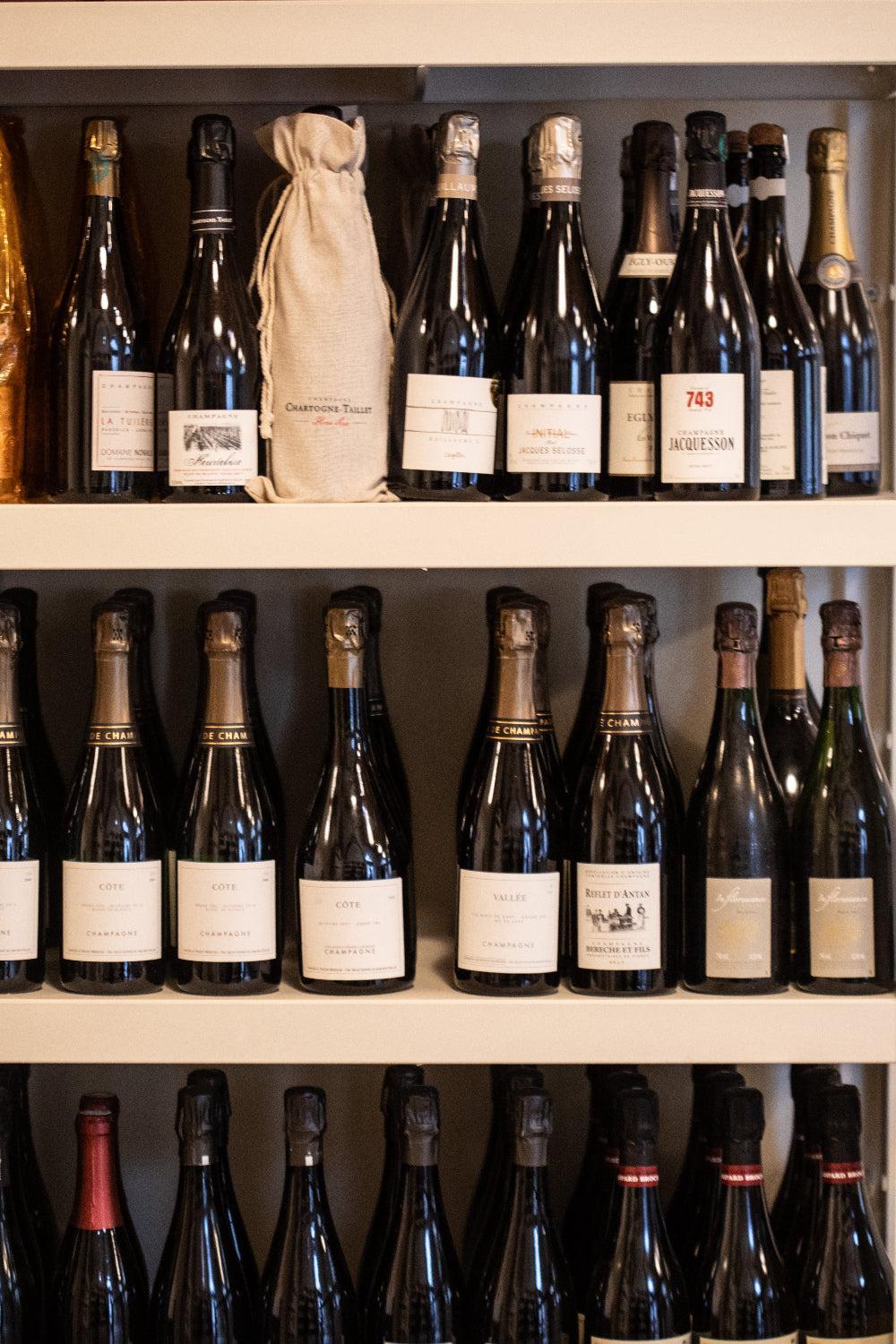 Rows of dark glass champagne bottles with elegant paper labels arranged on white wooden shelves in a cellar.