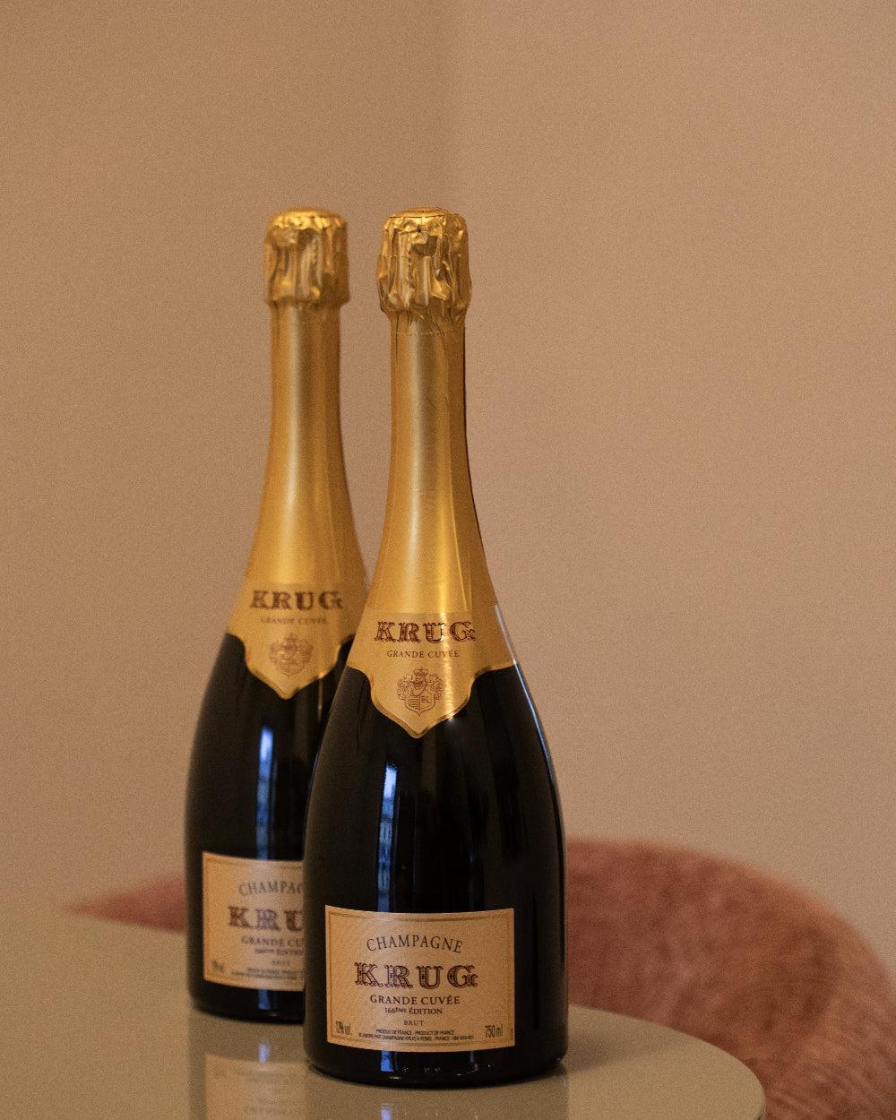 Two bottles of Krug Grande Cuvée champagne with gold foil tops standing on a reflective table against a neutral background.