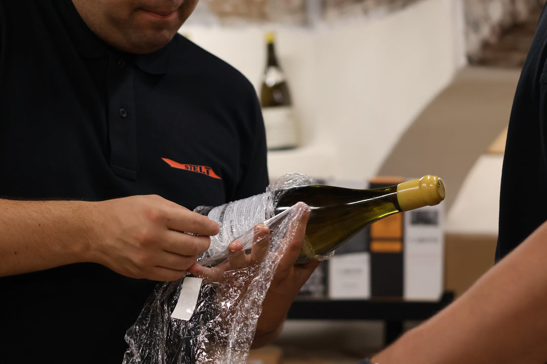Operatore in polo scura avvolge una bottiglia di vino in pellicola trasparente protettiva in un magazzino.