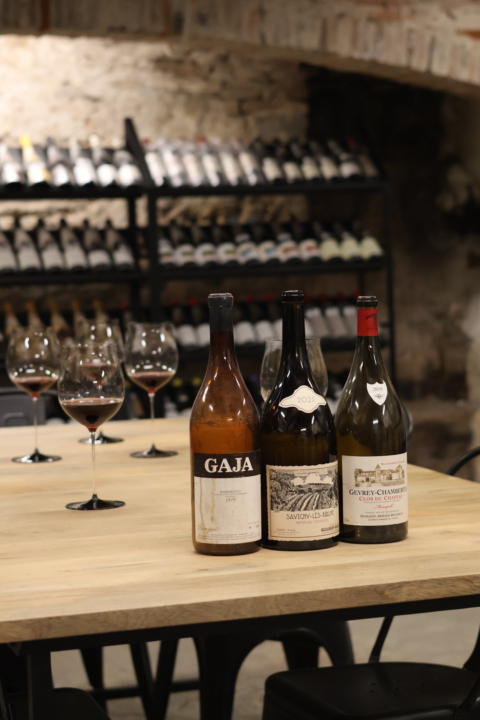 Three vintage wine bottles and crystal glasses on a wooden table in a rustic stone wine cellar with racks of bottles.