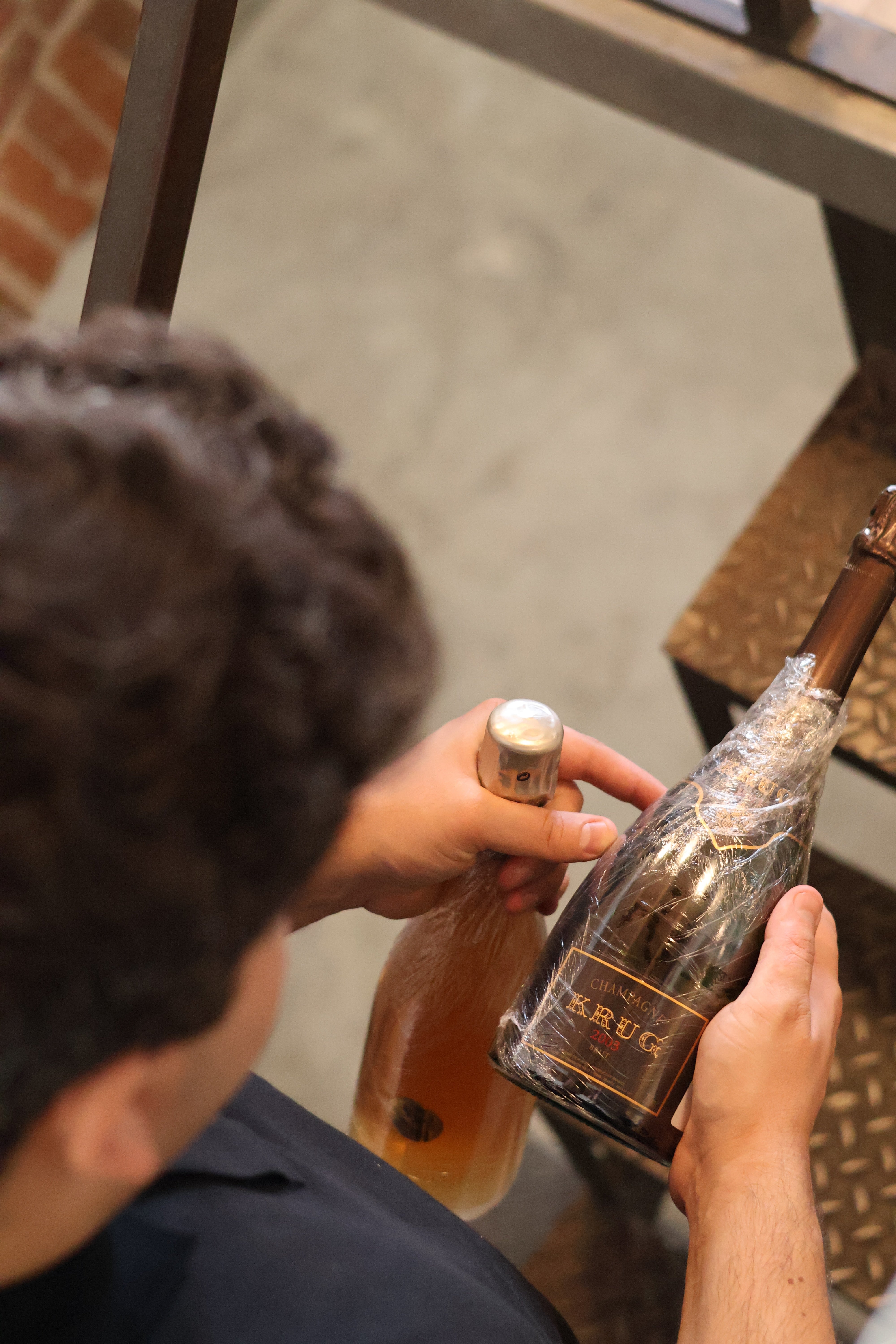 Persona che tiene in mano due bottiglie di Champagne Krug avvolte nella pellicola protettiva in un ambiente industriale.